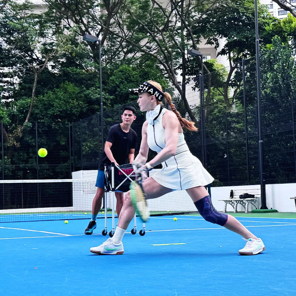 An adult player driving a two-handed backhand while a coach feeds from a ball cart
