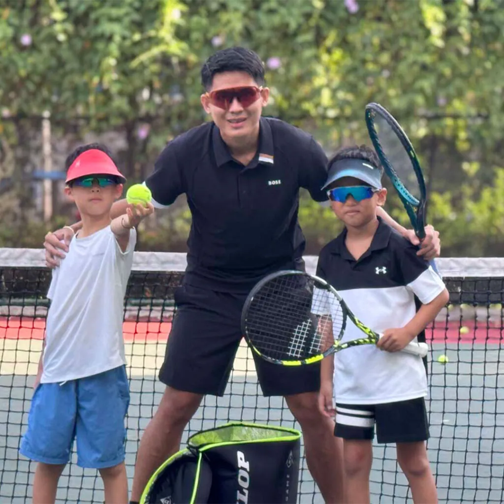 A coach on court with two young players holding racquets and a ball