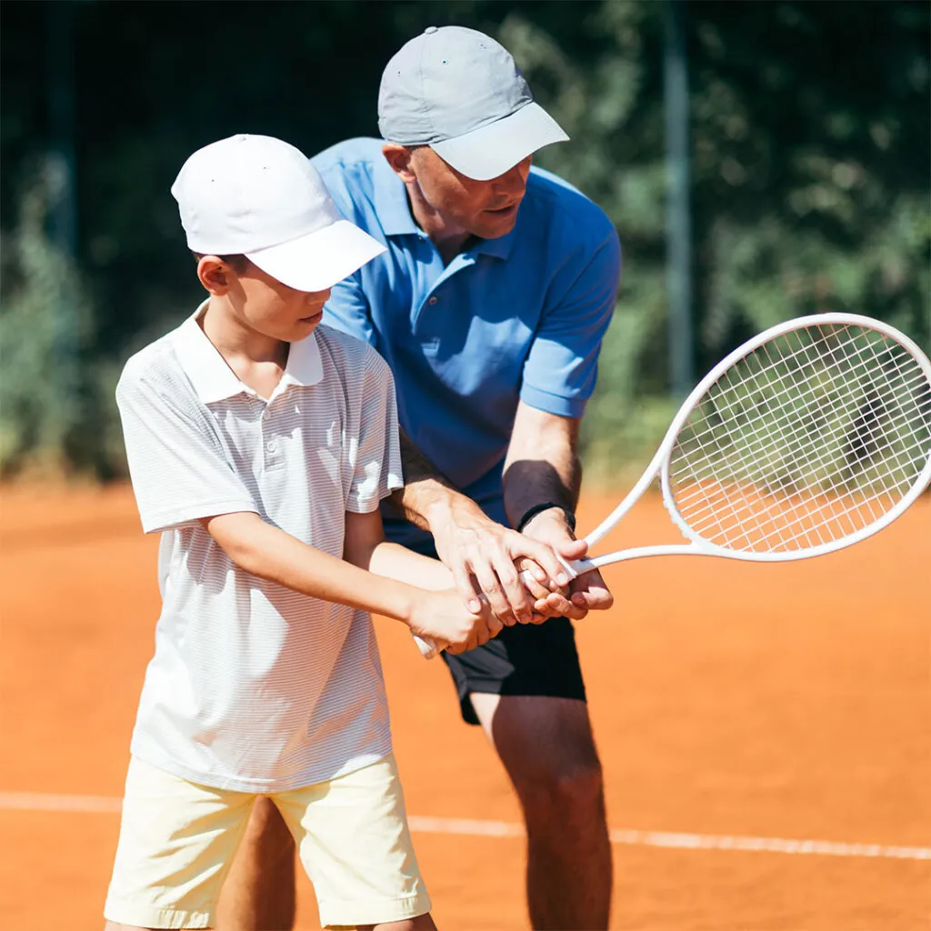 A coach shaping a young boy’s two-handed grip on a clay court