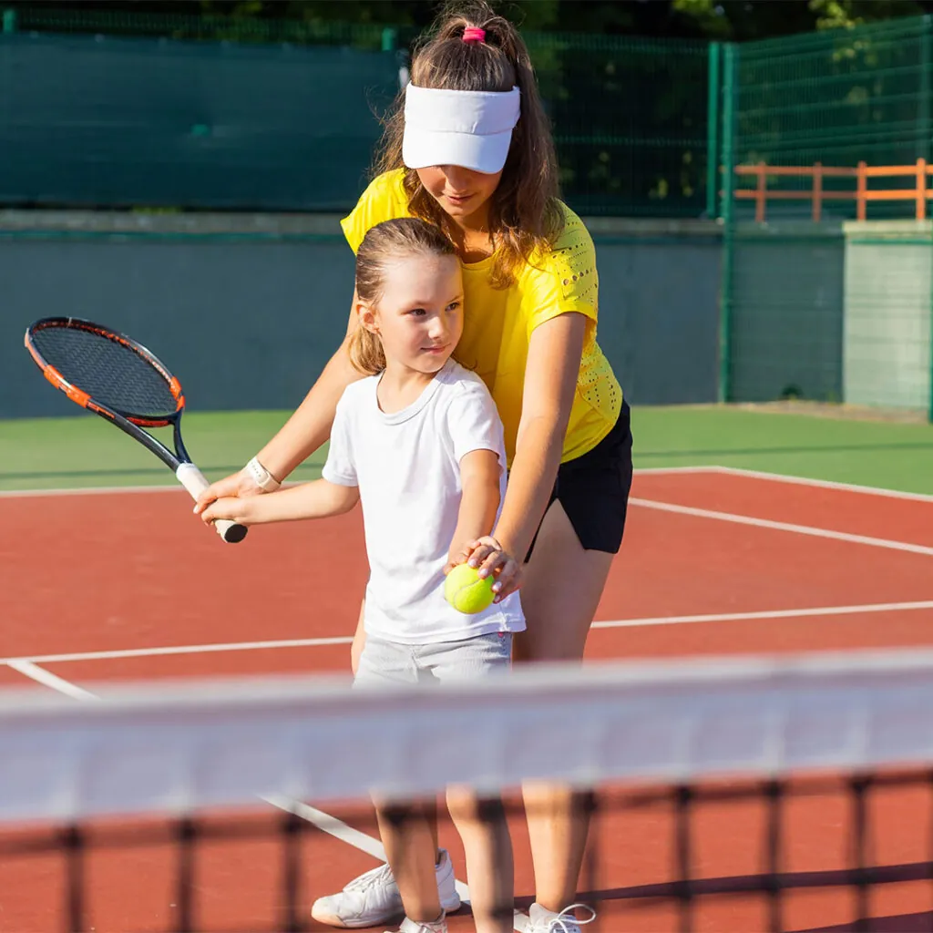 A coach standing behind a small child, guiding her racquet through a forehand
