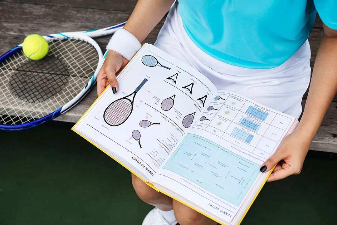 A player sitting courtside with a racquet, reading an illustrated guide to the racquet and the court