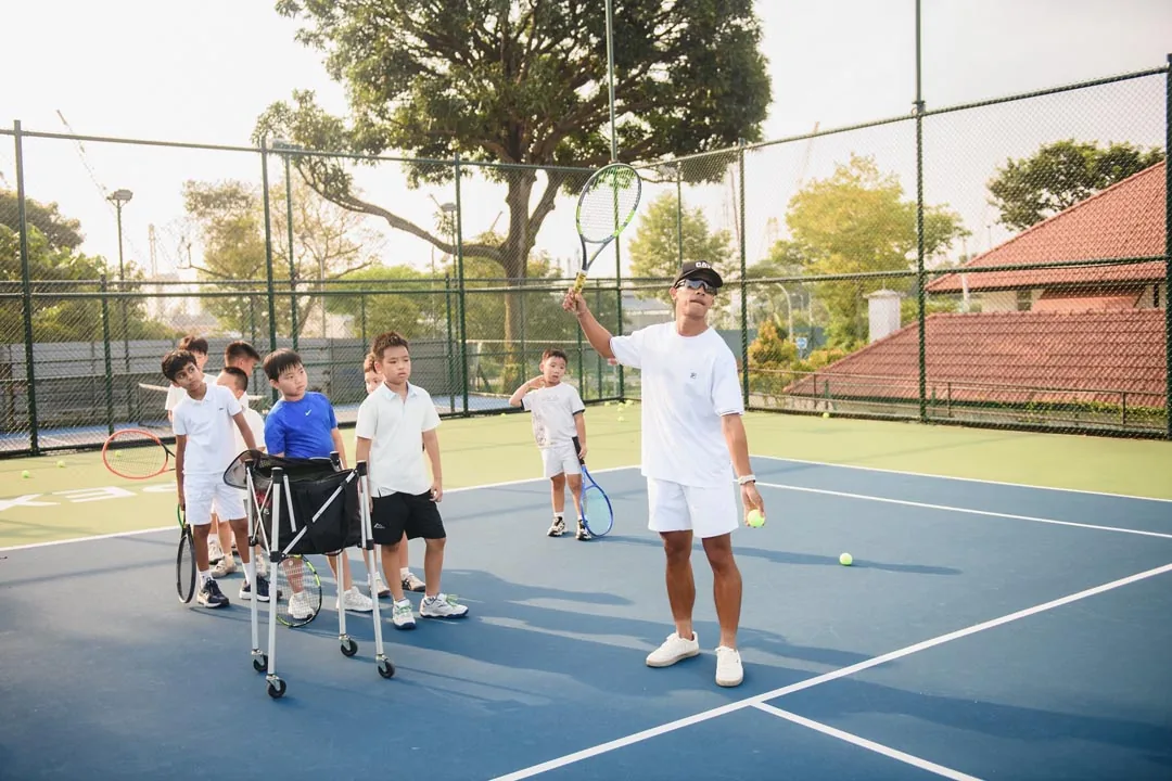 A coach with his arm raised, juniors lined up behind him holding racquets