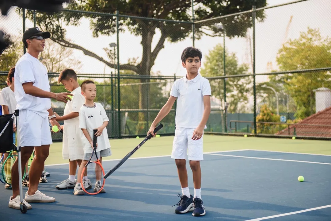 A coach feeding balls as a junior waits at the baseline with his racquet