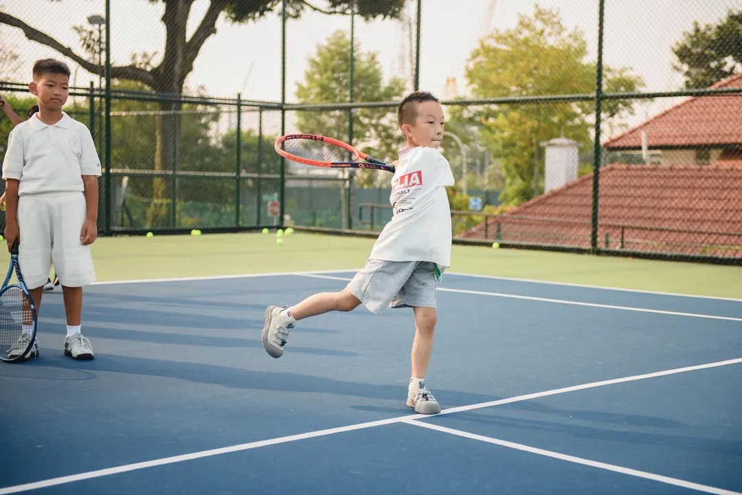 A boy following through on a forehand while a coach watches from behind
