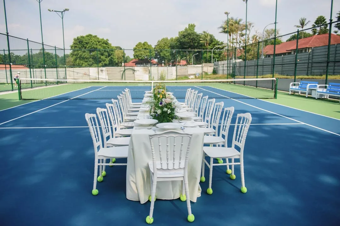 A long banquet table laid with white chairs and a floral centrepiece, set up on court